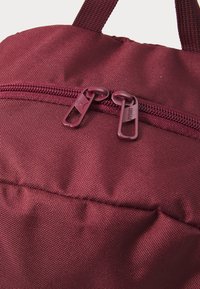 Burgundy reppu, jossa on teksturoitu kangas, varustettuna kaksoispurisilla muovivetoketjuilla ja yläkahvalla helppoa kantamista varten. Vetoketjuissa on logoyksityiskohta.