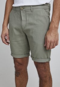 Pantalones cortos en verde oliva, hechos de un tejido texturizado. Presentan un cierre de botón, dobladillos enrollados y bolsillos laterales. Vista en primer plano del diseño.