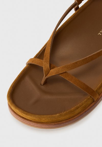 Bruna mockasin-sandaler med tunna remmar, en platt sula och en ljusbrun vadderad innersula. Designen har en minimalistisk stil och rena linjer.