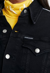 Svart denimjacka med silverfärgade tryckknappar, ett varumärkesmärke och kragdesign. Under finns en gul polotröja med vit text.