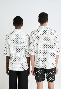 Krátke rukávy košele biele s čiernymi bodkami, skombinované s čiernymi šortkami, z pohľadu zozadu. Košele majú golier a voľný strih.