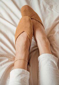 Bailarinas de ante en tono beige con un diseño de punta y un detalle recortado en la parte superior. Se llevan con pantalones blancos, apoyadas sobre una superficie de color claro.