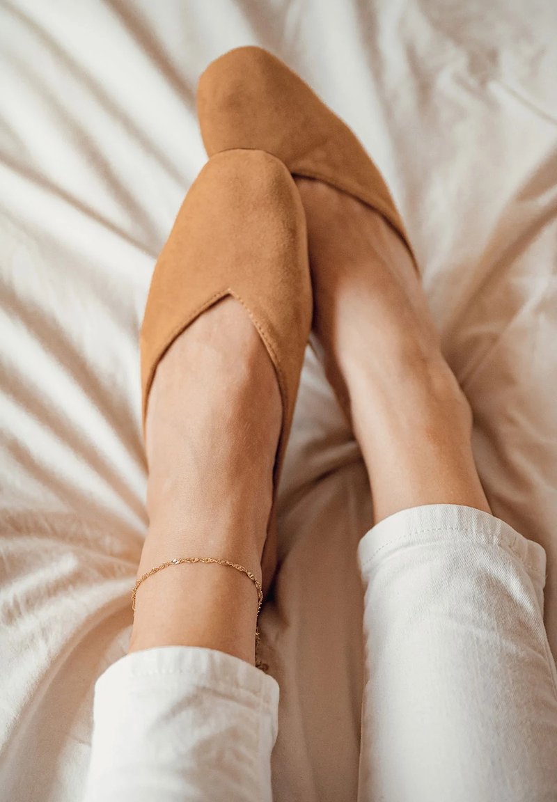 Bailarinas de ante en tono beige con un diseño de punta y un detalle recortado en la parte superior. Se llevan con pantalones blancos, apoyadas sobre una superficie de color claro.