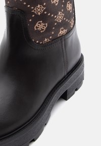 Bota de cuero marrón hasta el tobillo con parte superior de tejido con estampado, suela resistente con tacos y detalles de costura visibles. Textura suave en las partes de cuero.