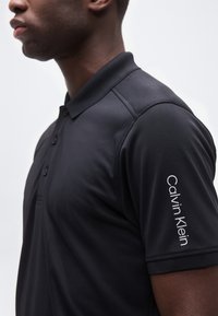 Άνδρας που φοράει μαύρο πόλο πουκάμισο Calvin Klein, με εμφανή τον γιακά, τα κουμπιά και το λογότυπο στο αριστερό μανίκι, μπροστά σε απλό φόντο.