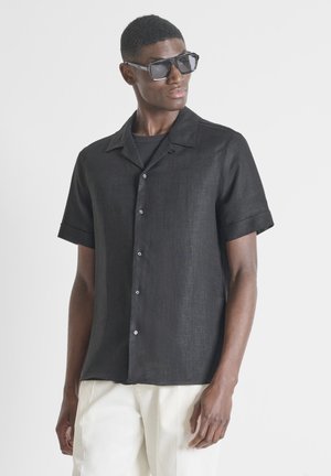 Man draagt een zwart shirt met korte mouwen en knopen, witte broek en grote donkere zonnebril, kijkt naar links tegen een effen achtergrond.