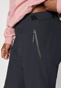 Μαύρο παντελόνι με πλαϊνή τσέπη με φερμουάρ, κατασκευασμένο από λείο, τεχνικό ύφασμα. Η μέση έχει κλείσιμο με αυτοκόλλητο για ρύθμιση του μεγέθους.