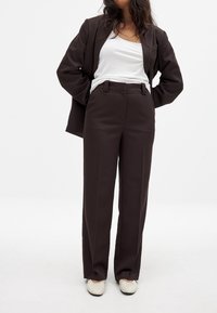Pantalons marron ajustés avec une coupe droite, poches latérales et passants de ceinture, associés à un débardeur blanc et un blazer assorti.