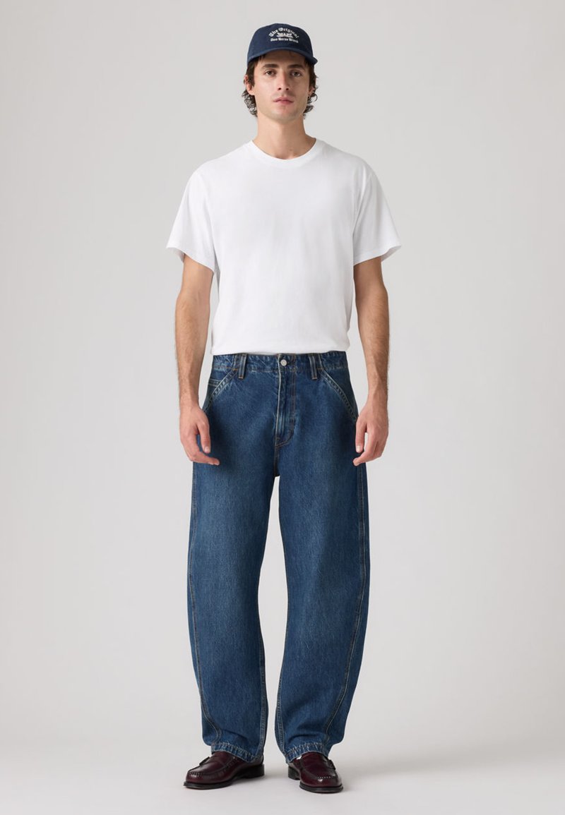 Camiseta blanca de cuello redondo, vaqueros azules de corte holgado y zapatos de cuero oscuro. Gorra con texto bordado. Fondo liso.