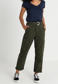 Pantalons cargo vert olive à taille haute, avec ceinture, plusieurs poches et ourlets retroussés, associés à un t-shirt bleu marine à col en V et des baskets noires.
