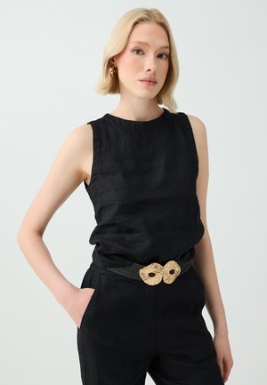 Femme blonde portant un haut noir sans manches et un pantalon noir avec une ceinture tissée comportant deux boucles beiges en forme de fleurs, debout avec une main dans la poche.