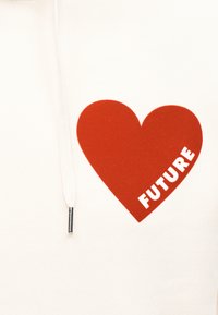 Tissu blanc avec un motif de cœur rouge sur la poitrine, contenant le mot "FUTURE" en lettres blanches en gras, et un cordon blanc avec embout en métal.