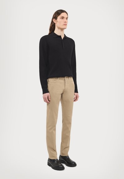 Crna rebrasta polo majica s dugim rukavima, bež slim-fit chinos hlače i crne kožne gležnjače, stojeći naslonjeni na običnu bijelu pozadinu.