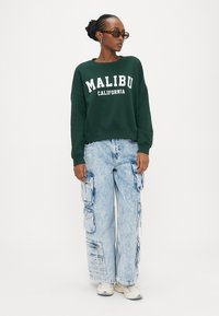 Grüner Sweatshirt mit weißer Aufschrift "MALIBU CALIFORNIA", kombiniert mit hellblauen Denim-Cargohosen mit mehreren Taschen und lockerem Schnitt.