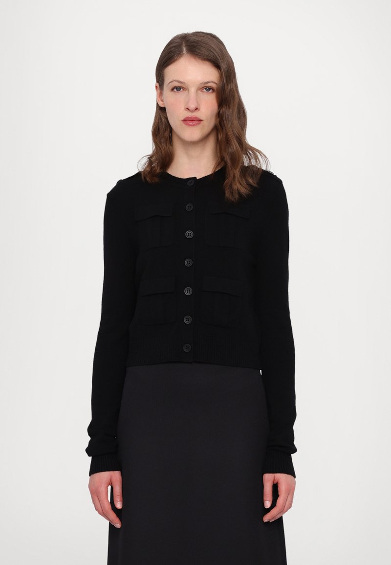 Cardigan nero lavorato a maglia con maniche lunghe, chiusura a bottoni e due tasche frontali. Polsini e orlo a coste, design corto, texture morbida.