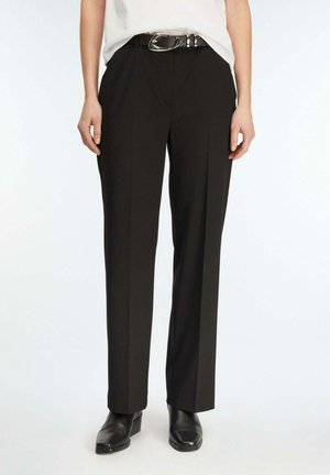 Personne portant un pantalon noir à jambes droites, une ceinture noire à boucle argentée, des bottes noires et une chemise claire rentrée.