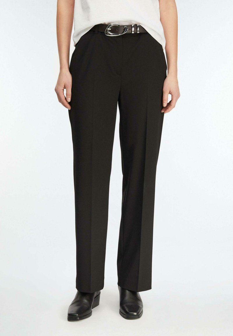 Persoon draagt zwart rechtvallende broek, zwarte riem met zilveren gesp, zwarte laarzen en een lichtgekleurd ingestopt overhemd.