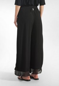 Mustat leveälahkeiset housut, joissa on rento istuvuus, joustava vyötärönauha ja läpikuultava kangashelma. Yhdistetty mustiin matalakantaisiin sandaaleihin.