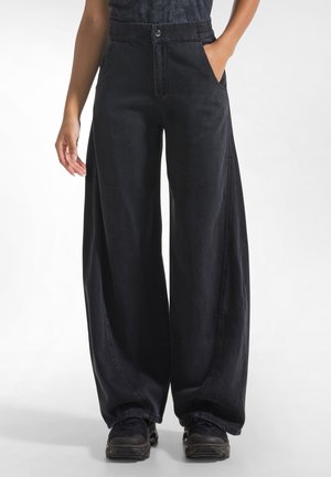 Persoon draagt zwarte broek met wijde pijpen en elastische tailleband, één hand in de zak, zwarte sneakers, staand tegen een lichte achtergrond.