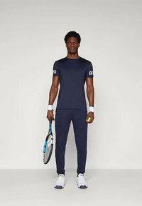 Marint atletisk outfit med en kortärmad topp och slim-fit byxor. Modellen håller en tennisracket och en boll, iklädd vita sneakers.