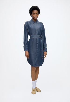 Kvinde står iført en langærmet denimkjole med knapper og bindebånd i taljen, hvide sokker og beige loafers på en ensartet hvid baggrund.