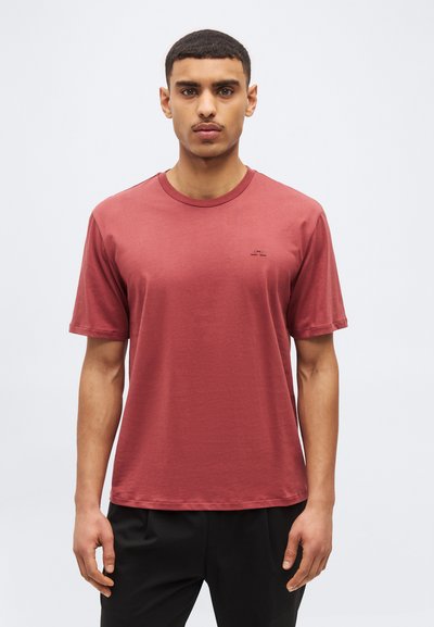 Junger Mann mit kurzen Haaren, der ein schlichtes, staubrosafarbenes T-Shirt mit kurzen Ärmeln und schwarze Hose vor weißem Hintergrund trägt.