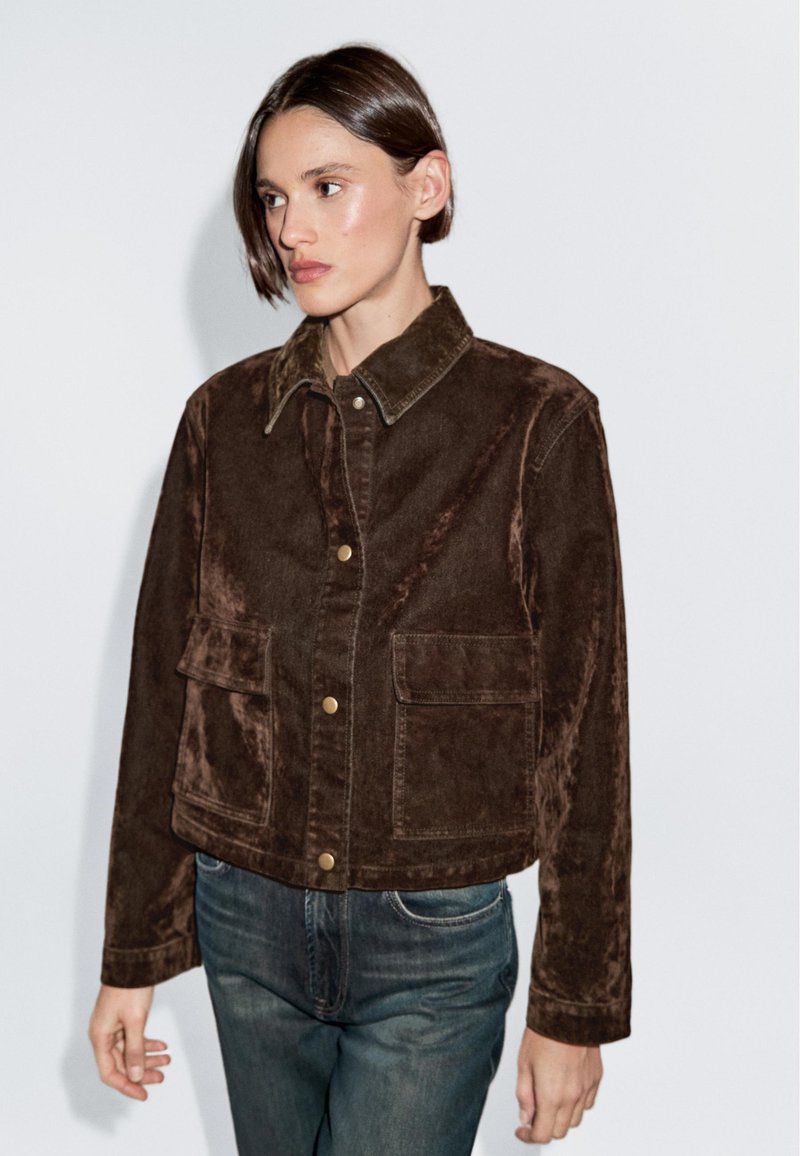 Vrouw met kort donker haar, gekleed in een donkerbruine corduroy jas met drukknoopjes en voorzakken, in combinatie met een blauwe spijkerbroek, staand tegen een witte muur.