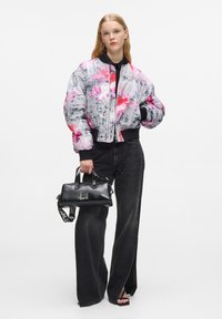 Giacca bomber colorata con una stampa astratta grigia, rosa e bianca, abbinata a pantaloni in denim neri a vita alta e una piccola borsa nera.