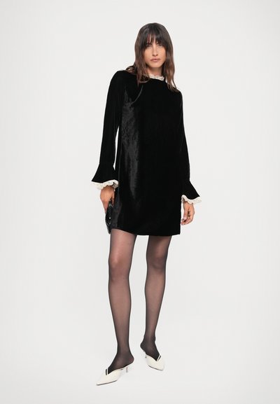 Black velvet dress with long bell sleeves, lace trim at the neckline and cuffs, worn with black tights and white heeled mules.