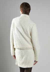 Hvid tekstureret jakke med en fuzzy stof, krave-design og en matchende nederdel. Kanterne har diskrete syninger. Bageste udsigt.