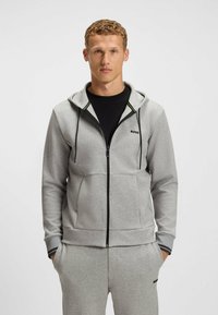 Felpa grigia con zip realizzata in tessuto morbido, dotata di cappuccio, con dettagli neri, tasche laterali e un piccolo logo sul petto.