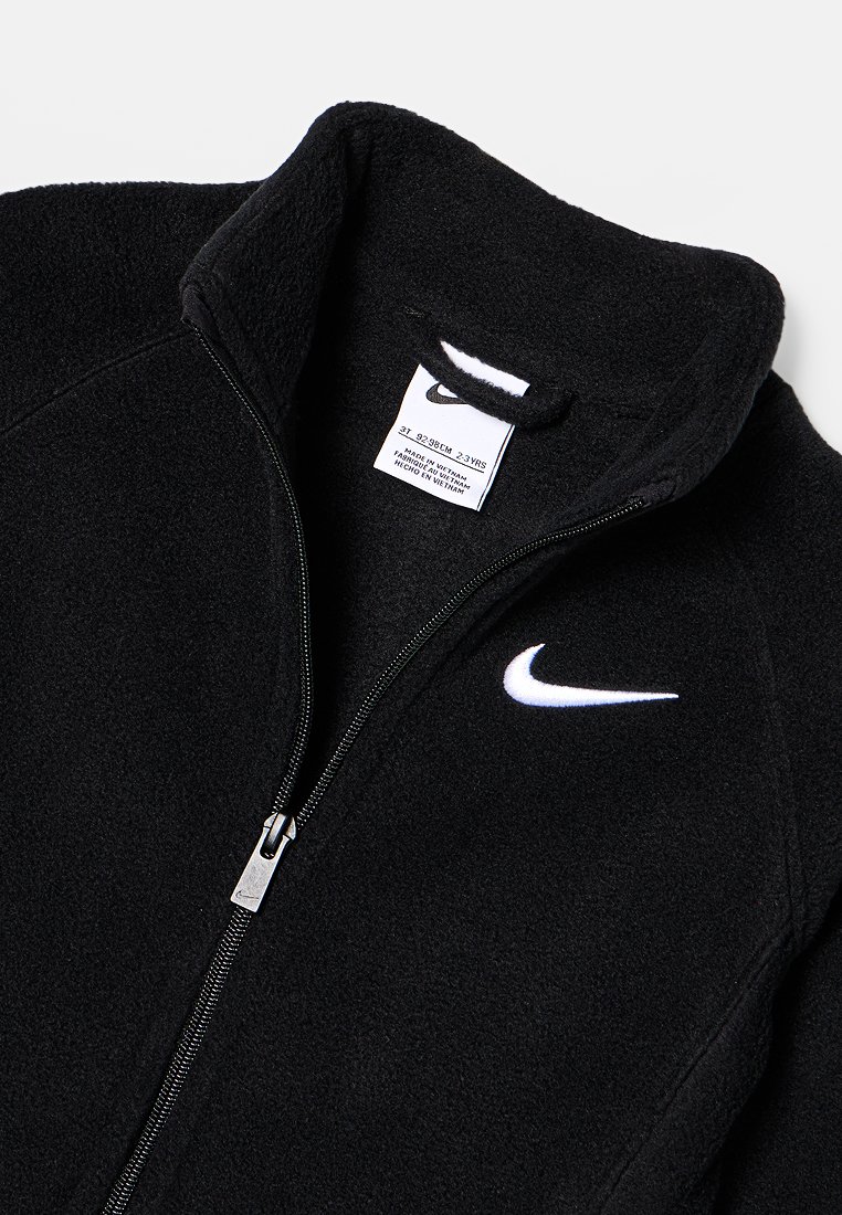 Schwarze Fleecejacke mit hohem Kragen, voller Reißverschluss und weißem Nike-Logo auf der Brust. Pflegehinweise im Inneren sichtbar.