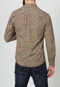Groen shirt met bloemendruk, voorzien van rode en blauwe bloemen. Lange mouwen, afgeronde zoom en een staande kraag. Gecombineerd met donkerblauwe jeans.