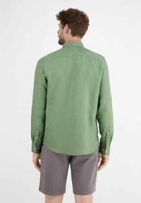 Groene langemouwen shirt van lichtgewicht stof; heeft een standaard kraag, knopenmanchetten en een iets gebogen zoom. Gecombineerd met grijze shorts.