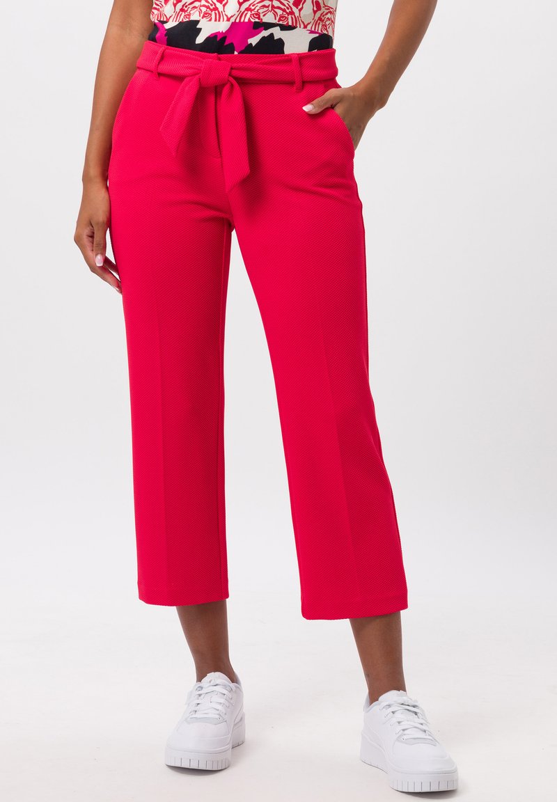 Bright pink cropped pants with a textured fabric, featuring a tied belt at the waist. Paired with white sneakers.