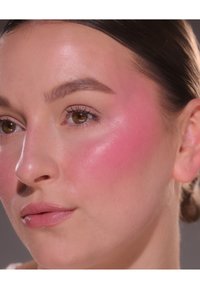 Nahaufnahme eines Gesichts mit glatter Haut und glänzendem Finish. Heller rosa Rouge betont die Wange. Braune Augen und eine helle, natürliche Lippenfarbe sind sichtbar.