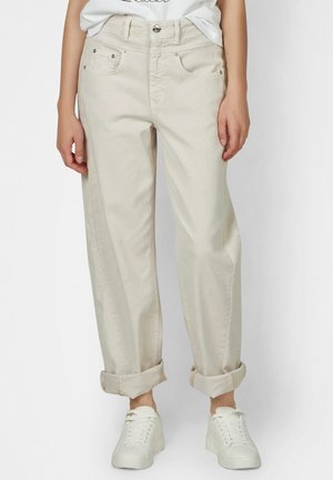 Person, die helle beige hochgekrempelte Hose und weiße Turnschuhe trägt und vor einem schlichten Hintergrund steht.