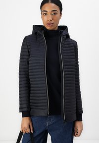 Veste matelassée noire avec capuche, dotée d'une fermeture éclair et de poignets côtelés. Portée sur un col roulé noir, associée à un jean bleu.