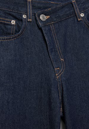 Gros plan sur le devant d'un jean en denim bleu foncé avec bouton métallique, braguette zippée, poches avant et détails de couture orange.