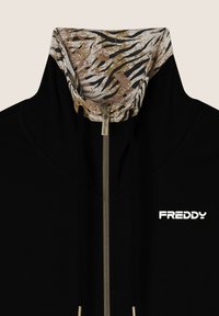 Sudadera con cremallera negra y un cuello de color marrón claro y negro con un patrón, que presenta una cremallera dorada y el logotipo blanco "FREDDY" en el pecho.