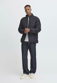 Sort pufferjakke med høj krave, frontlynlås, elastiske manchetter og sidelommer, båret over en hvid T-shirt og mørke jeans.