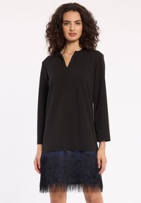 Schwarzes Langarmkleid mit V-Ausschnitt, das einen strukturierten blauen Fransenrand aufweist. Der Stoff wirkt glatt und leicht.