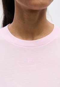 Κοντινό πλάνο ενός ατόμου που φοράει ανοιχτό ροζ μπλουζάκι με στρογγυλή λαιμόκοψη και μικρό ανάγλυφο λογότυπο "F" στο κέντρο του στήθους.