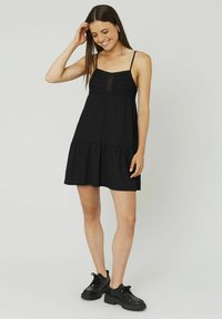 Robe noire sans manches avec un corsage smocké texturé, une jupe à volants et des bords festonnés, associée à des chaussures noires épaisses.