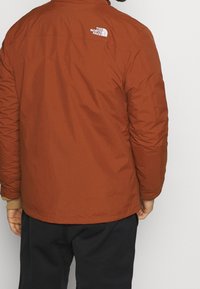 Zateplená bunda tmavě oranžové barvy s hladkým povrchem, bílým logem na zádech, elastickými manžetami a volným střihem.