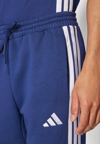 Jogging en bleu marine dotés d'un design à trois rayures blanches et d'un logo sur la jambe gauche. Comprend une taille élastique et un cordon de serrage.