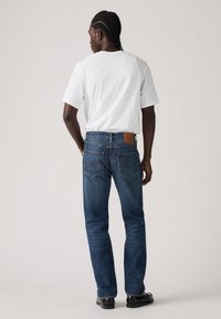 Blugi din denim albastru, cu croială dreaptă, având o etichetă din piele maro pe talie și cusături detaliate pe buzunare. Purtați cu un tricou alb.