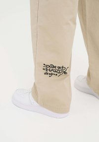 Beżowe spodnie o luźnym kroju, z czarnym haftowanym logo i akcentami. Połączone z białymi sneakersami, które mają gładką teksturę.