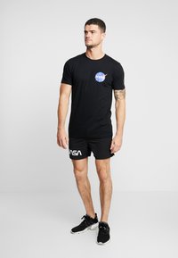Black cotton t-shirt with NASA logo in blue and red on the chest, paired with black shorts featuring white NASA text, worn with black sneakers.