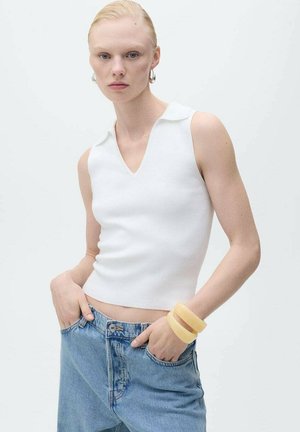 Haut sans manches blanc avec un col en V et une texture côtelée, porté avec un jean taille haute bleu clair. Le mannequin a les mains dans les poches, portant des bracelets jaune.